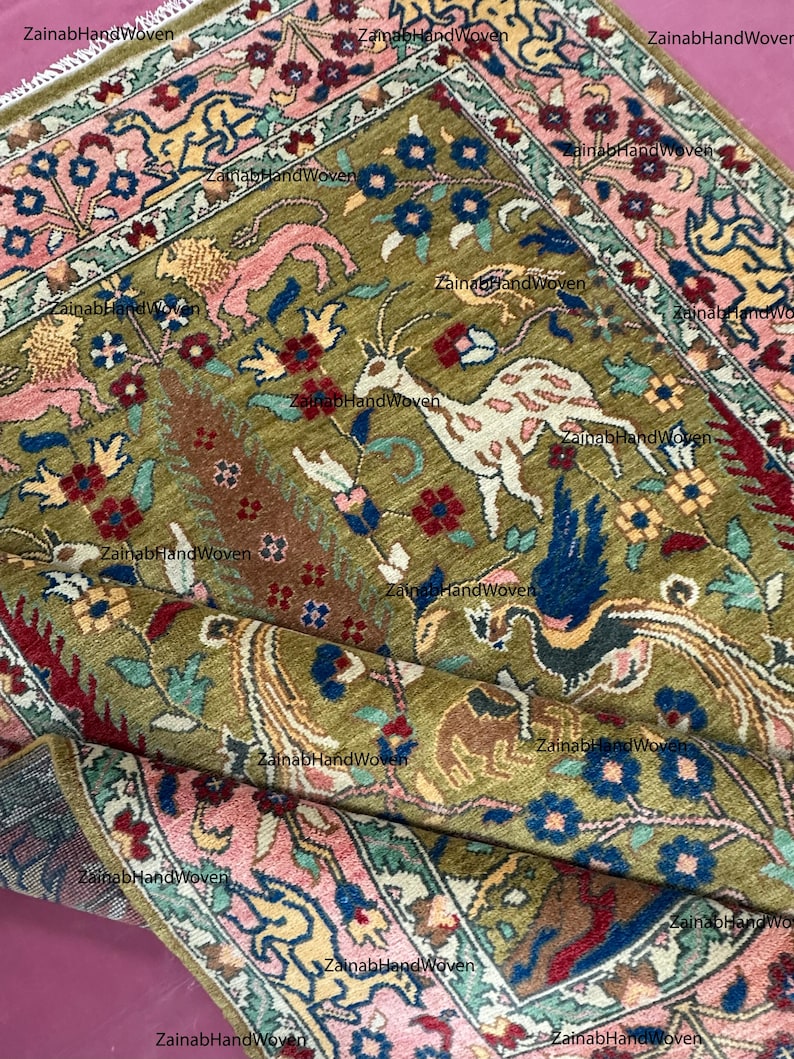 May include: An olive green, hand-woven rug with a detailed floral and animal pattern. The rug features a central design with deer and birds, surrounded by a border of pink, blue, and yellow floral motifs. The rug's dimensions are not visible.