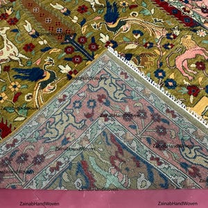 May include: An intricately designed rug with a central section in olive green, featuring animal and floral motifs in shades of white, blue, red, and pink. The rug's border is a contrasting pink with detailed patterns. The rug is hand-woven.