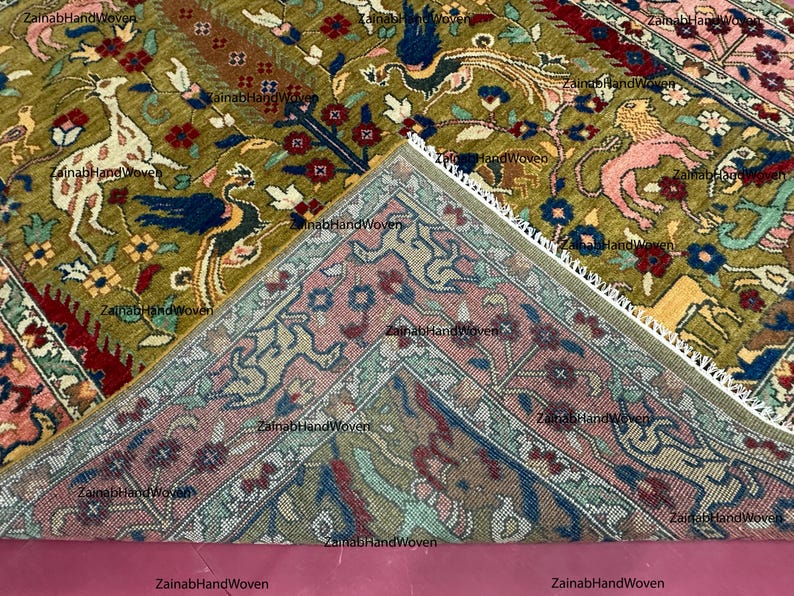 May include: An intricately designed rug with a central section in olive green, featuring animal and floral motifs in shades of white, blue, red, and pink. The rug's border is a contrasting pink with detailed patterns. The rug is hand-woven.