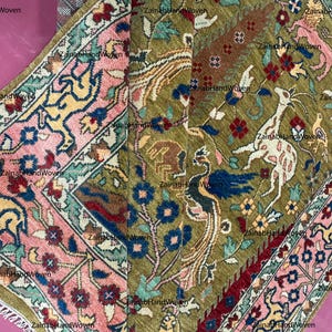 May include: Hand-woven rug with a detailed pattern. The rug features a central field of olive green with animal and floral motifs in white, blue, red, and brown. A pink and blue border frames the design. The rug is made of wool.