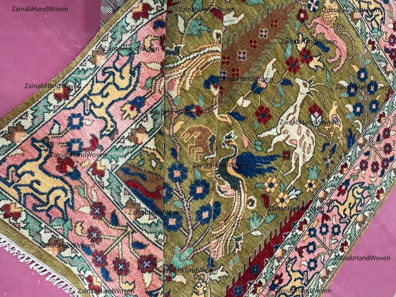 May include: Hand-woven rug with a detailed pattern. The rug features a central field of olive green with animal and floral motifs in white, blue, red, and brown. A pink and blue border frames the design. The rug is made of wool.