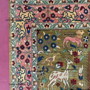 May include: A section of a hand-woven rug with a detailed floral and animal design. The rug features a central field of olive green with pink, blue, and yellow accents. The border is a mix of pink and green, with a fringe edge.