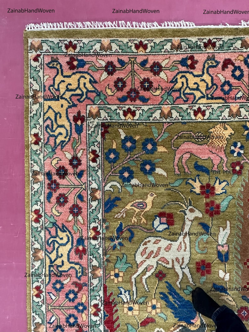May include: A section of a hand-woven rug with a detailed floral and animal design. The rug features a central field of olive green with pink, blue, and yellow accents. The border is a mix of pink and green, with a fringe edge.