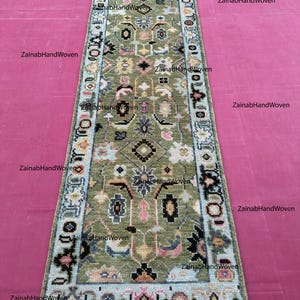 May include: A rectangular, hand-woven rug with a central olive green field and intricate floral and geometric patterns in various colors. The border is light blue with decorative elements. The rug has a fringe on one end.