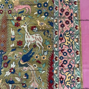 May include: Close-up of a section of a hand-woven rug with a detailed floral and animal design. The rug features a central olive green field with deer, birds, and floral motifs. The border is pink and green with more floral and animal patterns. The text "Zainab HandWoven" is visible.