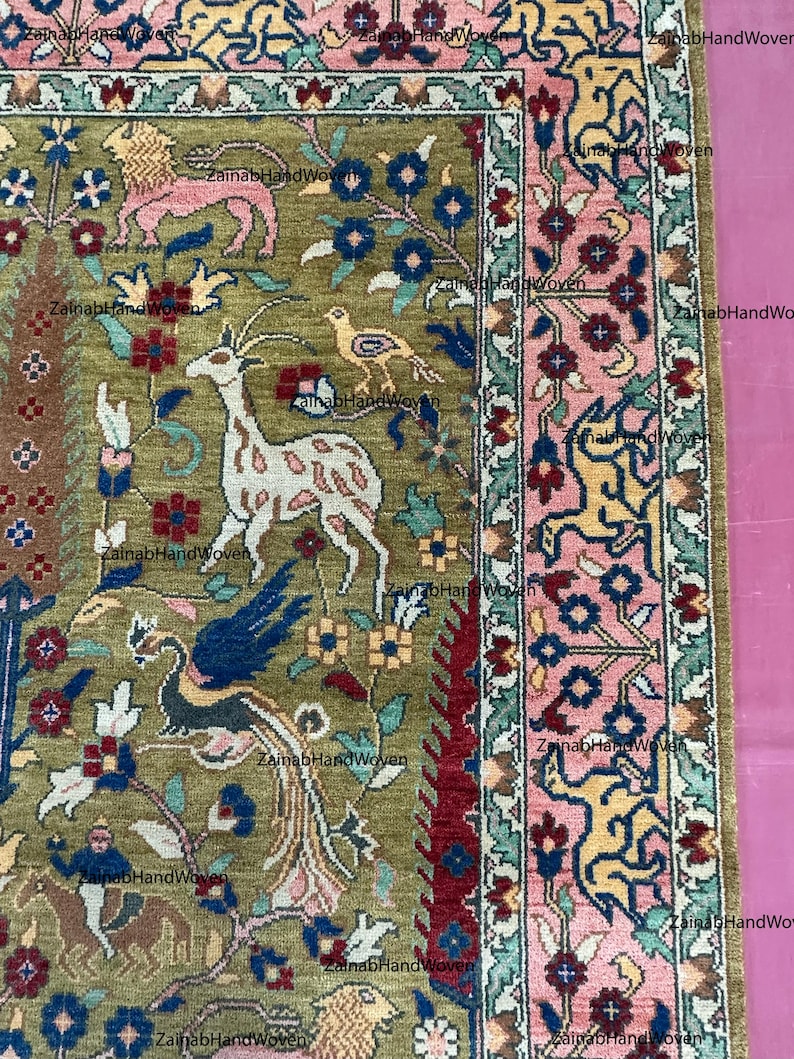 May include: Close-up of a section of a hand-woven rug with a detailed floral and animal design. The rug features a central olive green field with deer, birds, and floral motifs. The border is pink and green with more floral and animal patterns. The text "Zainab HandWoven" is visible.