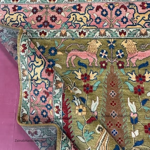 May include: An olive green and pink hand-woven rug with a detailed floral and animal pattern. The rug features a border with stylized animals and floral motifs, with a central design of trees and flowers. The rug is folded, revealing the intricate design.