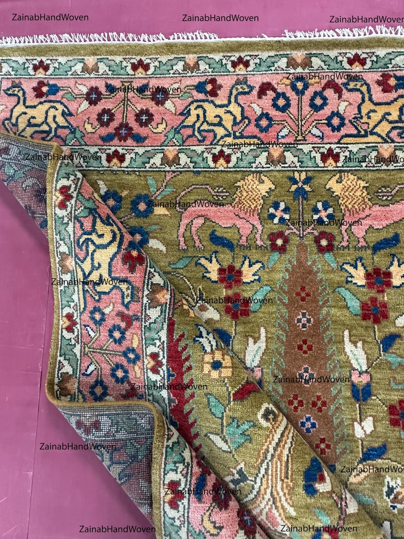 May include: An olive green and pink hand-woven rug with a detailed floral and animal pattern. The rug features a border with stylized animals and floral motifs, with a central design of trees and flowers. The rug is folded, revealing the intricate design.