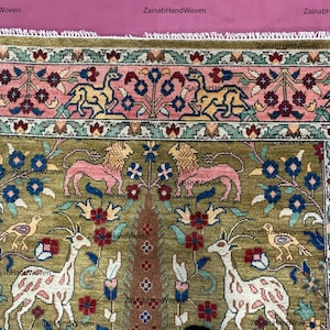 May include: Hand-woven rug with a detailed floral and animal design. The rug features a central olive green field with pink lions, deer, and birds. The border is pink with floral patterns, and the rug has a fringed edge. The text "ZainabHandWoven" is visible.