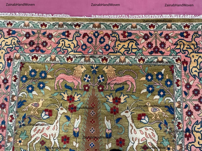 May include: Hand-woven rug with a detailed floral and animal design. The rug features a central olive green field with pink lions, deer, and birds. The border is pink with floral patterns, and the rug has a fringed edge. The text "ZainabHandWoven" is visible.