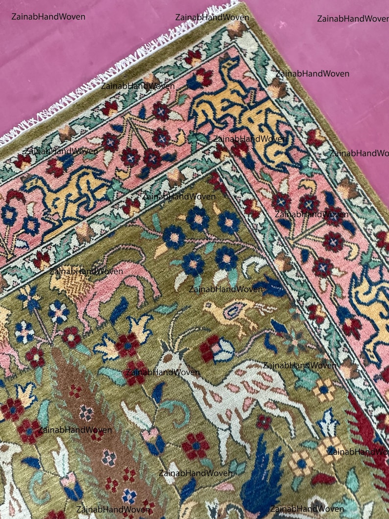 May include: A detailed, hand-woven rug with a central olive green field featuring animal and floral motifs in pink, blue, and white. The border is a contrasting pink with yellow and blue stylized designs. The rug has a fringed edge.