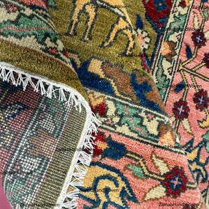 May include: Close-up of a hand-woven rug with intricate patterns. The rug features a combination of colors, including pink, green, blue, and yellow. The design includes floral and animal motifs, with a fringed edge.