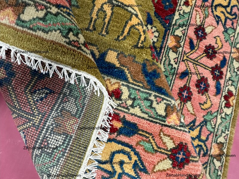 May include: Close-up of a hand-woven rug with intricate patterns. The rug features a combination of colors, including pink, green, blue, and yellow. The design includes floral and animal motifs, with a fringed edge.