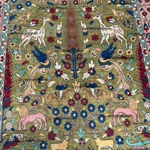 May include: An intricately designed rug with a central olive green field, featuring stylized animals, birds, and floral motifs in pink, blue, and brown. The rug has a detailed border with a pink and red pattern, showcasing a traditional design.