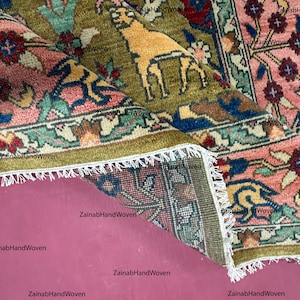 May include: Close-up of a folded, hand-woven rug with a detailed floral and animal pattern. The rug features a central field of olive green with a golden animal figure, surrounded by pink, blue, and red floral borders. White fringe edges the rug.