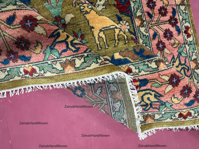 May include: Close-up of a folded, hand-woven rug with a detailed floral and animal pattern. The rug features a central field of olive green with a golden animal figure, surrounded by pink, blue, and red floral borders. White fringe edges the rug.