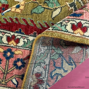 May include: Close-up of a hand-woven rug with intricate floral patterns. The rug features a combination of colors, including red, green, pink, and blue. The edges are trimmed with a golden border. The rug is partially rolled up, revealing the underside.