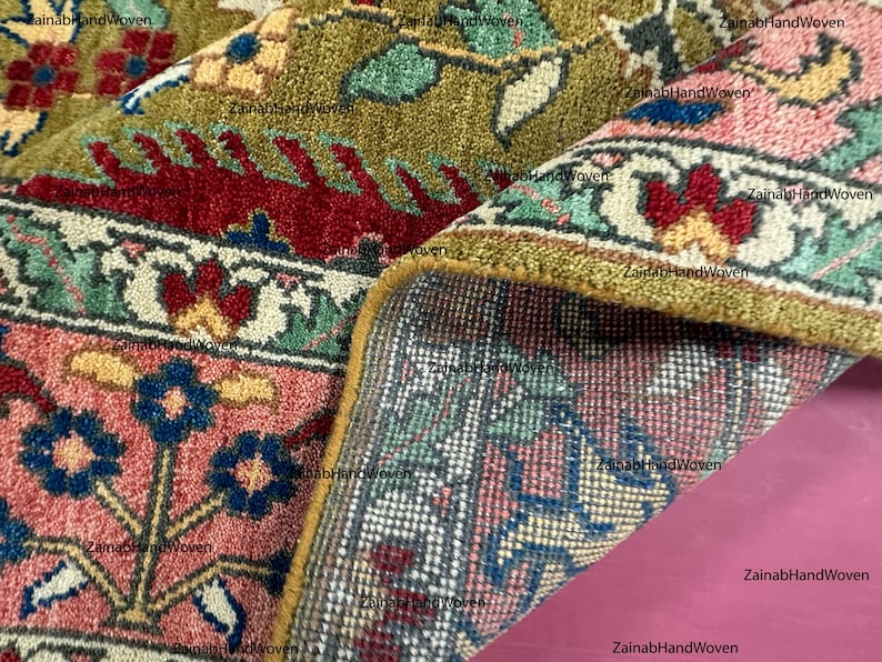 May include: Close-up of a hand-woven rug with intricate floral patterns. The rug features a combination of colors, including red, green, pink, and blue. The edges are trimmed with a golden border. The rug is partially rolled up, revealing the underside.