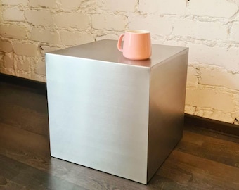 May include: A brushed metal cube-shaped side table with a pink mug on top. The table is a light silver color and sits on a dark wood floor. The background is a white brick wall.