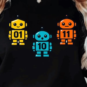 Peut inclure: Sweat-shirt noir avec trois robots colorés : jaune, bleu et orange. Chaque robot porte un numéro : 01, 10 et 11. Le texte 'Digitale Product Download' est visible. Produit numérique.