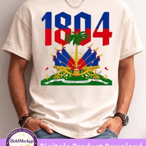 Peut inclure: T-shirt crème avec l'année "1804" en rouge et bleu, et un design aux armoiries haïtiennes. Le design comprend des drapeaux, des canons et le texte "UNION FAIT LA FORCE".