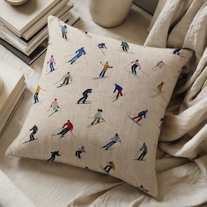 Può includere: Cuscino quadrato beige con sciatori ricamati in vari abiti colorati. Gli sciatori sono raffigurati in azione, con sci e bastoncini, su uno sfondo neutro. Il cuscino è posto su un letto con una coperta colorata neutra.