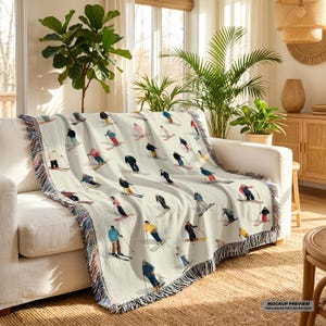 May include: Cream-coloured woven throw blanket featuring a repeating pattern of skiers in various colourful outfits. The blanket has a fringed edge with a mix of colours. The blanket is draped over a white sofa.