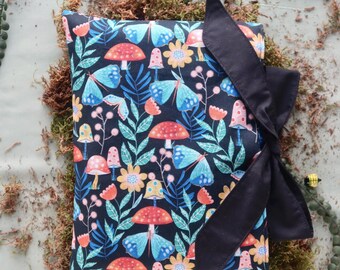 May include: A fabric-covered book or journal featuring a whimsical pattern of mushrooms, butterflies, and flowers in shades of red, blue, and yellow on a dark background. A black ribbon is tied around the journal.