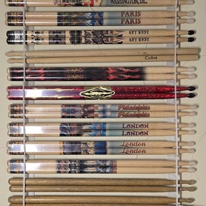 Pode incluir: Uma coleção de baquetas de bateria exibidas em um rack montado na parede. As baquetas apresentam vários designs e nomes de cidades, incluindo "Miami", "Washington, D.C.", "Paris", "Key West", "Denver" e "Boston".