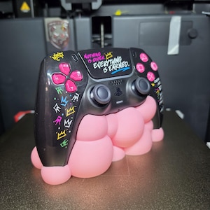 May include: A black gaming controller featuring pink buttons and colorful crown graphics on the left side. The center panel displays the white text "NOTHING IS GIVEN. EVERYTHING IS EARNED." The controller rests on a pink, bubble-textured stand.