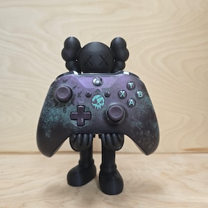 May include: A black figurine holding a purple and black Xbox controller. The controller has a skull and crossbones design and the letters X, Y, A, and B. The figurine has a round head with two large ear-like shapes and crossed-out eyes.