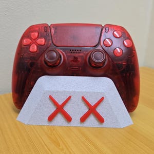 Könnte beinhalten: Ein transparent roter Videospiel-Controller mit roten Knöpfen und einem grauen Ständer mit zwei roten "X"-Symbolen. Der Controller ruht auf einem weiß-grauen Ständer.