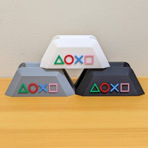 May include: Three 3D-printed controller button-shaped stands in white, gray, and black. Each stand features the green triangle, red circle, blue X, and pink square symbols. The stands are displayed on a wooden surface.