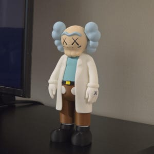 May include: A figurine of a cartoon character with a light tan face, blue hair, and crossed-out eyes. The figure wears a white lab coat, a light blue shirt, brown pants, and black shoes. The figure is standing on a black surface.