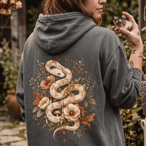 Könnte beinhalten: Ein anthrazitfarbener Kapuzenpullover mit einer detaillierten Grafik auf der Rückseite, die eine geringelte cremefarbene Schlange umgeben von orangefarbenen und weißen Blumen, grünen Blättern und kleinen Pilzen zeigt. Das Motiv ist mittig platziert.
