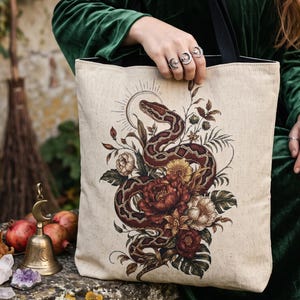 Pode incluir: Uma sacola de lona bege com uma ilustração detalhada de uma cobra enrolada entrelaçada com flores vermelho-escuras e brancas, folhas verdes e um motivo de sol. A bolsa tem alças pretas e um formato retangular com base plana.