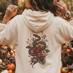 May include: A cream-coloured hooded sweatshirt featuring a detailed graphic on the back. The design depicts a coiled snake intertwined with various flowers, including a large red rose, surrounded by green leaves and thin, radiating sunburst lines.
