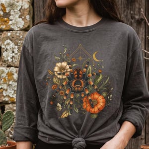 Può includere: Una maglietta a maniche lunghe grigio antracite con una grafica centrale di una tarantola circondata da fiori arancioni, fogliame verde, una luna crescente e una zucca. La maglietta ha un girocollo e un nodo sull'orlo anteriore.
