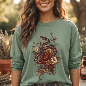 Op de afbeelding: Een saliegroen shirt met lange mouwen en een centrale grafische print van een kronkelende slang verweven met diverse bloemen en bladeren in rood-, crème- en bruintinten, gecombineerd met een versleten blauwe spijkerbroekshort.