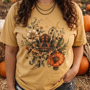 May include: A mustard yellow short-sleeve t-shirt featuring a graphic print of a large brown and black tarantula surrounded by orange flowers, green foliage, a small crescent moon, and geometric line art on the front center.