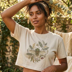 May include: A cream-colored short-sleeved crop top featuring a central graphic print of a green beetle surrounded by botanical illustrations of ferns, leaves, and small flowers in muted green, yellow, and brown tones.