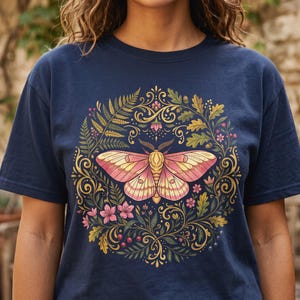Op de afbeelding: Een marineblauw t-shirt met een centrale afbeelding van een roze en gele nachtvlinder, omringd door een cirkelvormige krans van groene varenbladeren, eikenbladeren, kleine roze bloemen en rode bessen met gouden, krullende decoratieve accenten.
