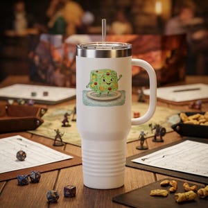 Könnte beinhalten: Weißer Reisebecher mit silbernem Deckel und Griff. Der Becher zeigt eine Cartoon-Illustration eines lächelnden grünen Gelee-Desserts mit Früchten. Ein klarer Strohhalm erstreckt sich vom Deckel. Der Becher steht auf einem Holztisch mit Spielsteinen.