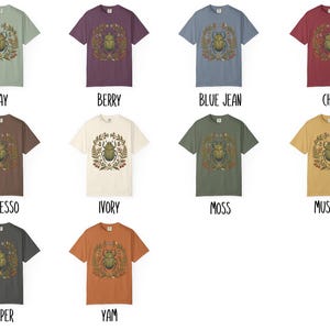 K&ouml;nnte beinhalten: Eine Auswahl an kurz&auml;rmeligen T-Shirts in verschiedenen Farben, jedes mit einer zentralen Grafik eines Skarab&auml;us-K&auml;fers, der von einem botanischen Kranz umgeben ist. Die Shirts sind in Farbt&ouml;nen wie Bay, Berry, Blue Jean, Chili, Espresso, Ivory, Moss, Mustard, Pepper und Yam erh&auml;ltlich.