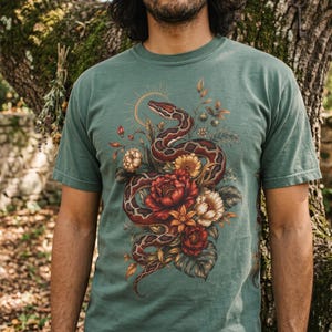 Op de afbeelding: Een saliegroen t-shirt met een gedetailleerde grafische print van een kronkelende slang verstrengeld met levendige rode, oranje en witte bloemen en bladeren. Het ontwerp bevat een dunne zonnestraal-omtrek boven de kop van de slang op de borst.