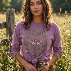 May include: A long-sleeve mauve t-shirt featuring a central graphic of a moth surrounded by a circular wreath of green leaves, gold vines, and small flowers. The shirt is tied in a knot at the waist and has a crew neckline with long sleeves.