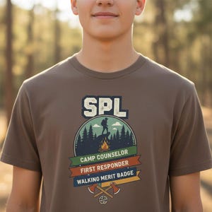 May include: A brown t-shirt featuring the text "SPL" in a white and blue block font. Below the text is a graphic of a person hiking on a mountain, with the words "CAMP COUNSELOR", "FIRST RESPONDER", and "WALKING MERIT BADGE".