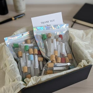 May include: A black box filled with clear test tubes with colored caps, packaged in holographic zip bags. A card with the words "THANK YOU" is visible. The box is lined with tissue paper.