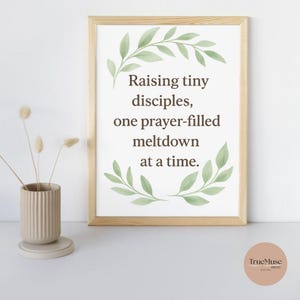 May include: A framed print with the text "Raising tiny disciples, one prayer-filled meltdown at a time." The print is surrounded by a light brown frame and green leaf accents. A beige vase with dried flowers is on the left.