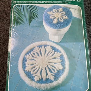 May include: A Bucilla Latch Hook Kit featuring a "Snowflake" Bath Ensemble. The kit includes a round mat and matching lid cover, both with a blue background and a white snowflake design. The mat is 24 inches in diameter. The kit contains premium acrylic yarn.
